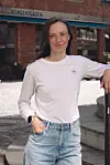 Kvinne i hvit genser og jeans står utendørs foran en bygning.