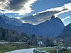 Fjellandskap med en bratt fjelltopp, skog, hus og en vei under blå himmel.