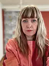 Portrett av en kvinne med langt lyst hår og rødrosa genser innendørs.