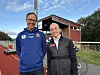 En kvinne og en mann poserer ved en friidrettsbane med løpebaner og klubbhus i bakgrunnen.