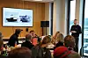 En mann står foran en sittende forsamling i et møterom, med bilder av ferger på en skjerm bak seg.