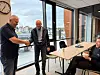 Tre menn møtes i et lyst kontor, der to av dem håndhilser mens en tredje sitter ved et bord.