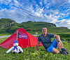 Mann sitter i gresset foran et rødt telt med en liten bamse, omgitt av grønne fjell under blå himmel.