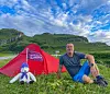 Mann sitter i gresset foran et rødt telt med en liten bamse, omgitt av grønne fjell under blå himmel.