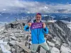En mann i blå jakke holder et lilla skilt på et steinete fjellplatå med snødekte fjell i bakgrunnen.