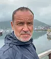 Mann i mørk jakke ved en havn med båter, fjord og fjell i bakgrunnen.