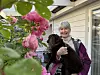 Gråhåret kvinne holder en svart hund ved inngangsdøren, med rosa roser i forgrunnen.