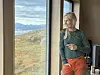 Kvinne står ved et vindu med fjellandskap og innsjø i bakgrunnen.
