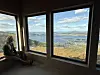 Person sitter ved store vinduer med utsikt over et fjordlandskap.