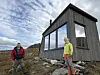 To turgåere står foran en trekledd hytte i et steinete fjellandskap.