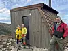 Tre turgåere står ved en liten trehytte i fjellet under en blå, skyet himmel.