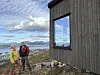 To turgåere står ved en mørk trekledd bygning foran et fjordlandskap med fjell.
