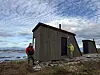 To personer ved et værslitt naust på en steinete kyst med sjø og skyer i bakgrunnen.