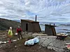 To personer ved små, værbitte hytter på et steinete svaberg med fjord i bakgrunnen.