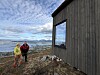 To turgåere står ved en trekledd hytte med hav og fjell i bakgrunnen.