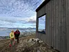 To turgåere står ved en trekledd hytte med hav og fjell i bakgrunnen.