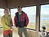 To personer står i en trebekledd hytte med store vinduer og utsikt over fjord og fjell.