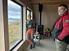To personer inne i en liten trehytte med vedovn og utsikt mot fjord og fjell.