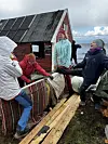 Flere personer i turklær arbeider ved en eldre rød trehytte med plank og tekstiler rundt seg.