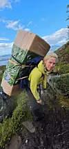 Turgåer med stor innpakket sekk klatrer opp en bratt fjellsti med fjord og fjell i bakgrunnen.