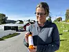 Kvinne med briller holder en flaske med oransje innhold utendørs ved en gård.