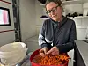 En person sorterer oransje tindvedbær i en rød kasse innendørs.
