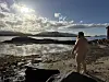 Naken person står på en steinete strand ved fjorden i sollys, med fjell i bakgrunnen.