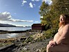 Naken person sitter på en steinete strand ved sjøen, med et rødt naust og skog i bakgrunnen.