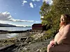 Naken person sitter på en steinete strand ved sjøen, med et rødt naust og skog i bakgrunnen.