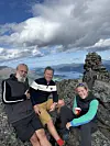 Tre personer poserer ved en steinvarde på et fjell med fjord og fjellandskap i bakgrunnen.