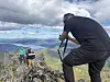 En person filmer fra en fjelltopp, mens to andre står på en steinrygg med fjord og fjell i bakgrunnen.