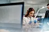 Laboratoriearbeider med blå hansker håndterer prøver bak en uskarp dataskjerm.