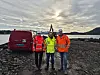 Tre arbeidere i synlighetsklær står ved en rød varebil på en steinete strand.