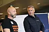 To menn står sammen på et innendørs arrangement foran en informasjonsvegg.