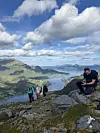 Fire turgåere på et steinete fjell med utsikt over en fjord, grønne fjellsider og blå himmel.