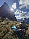 To fjellvandrere hviler i lyngen under en bratt fjelltopp i solskinn.