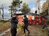To personer utenfor en rød hytte i skogen, med vann og fjell i bakgrunnen.