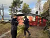 To personer utenfor en rød hytte i skogen, med vann og fjell i bakgrunnen.