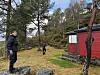 To personer ved en rød hytte i et skogkledd landskap.