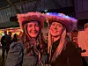 To smilende kvinner i fargerike fjærhatter på et lyst innendørs arrangement.