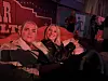 To kvinner sitter og smiler foran en scene med rødt sceneteppe.