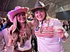 To smilende kvinner i cowboyhatter holder drikke under et innendørs arrangement.
