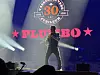 Vokalist synger på en konsertscene foran Plumbo-logoen og sterke scenelys.