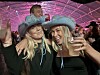 To kvinner i turkise cowboyhatter smiler på en fest med rosa lys og mennesker i bakgrunnen.