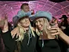To kvinner i turkise cowboyhatter smiler på en fest med rosa lys og mennesker i bakgrunnen.