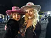 To kvinner i cowboyhatter poserer sammen i et opplyst arrangementslokale.