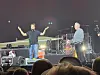 To musikere står på en opplyst scene foran publikum under en konsert.