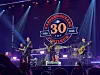 Band spiller på en opplyst scene foran publikum med en stor 30-årslogo i bakgrunnen.