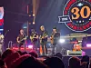 Band spiller på scene foran et publikum, med et stort 30-årsjubileumsskilt i bakgrunnen.