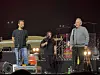 Tre musikere opptrer på en scene med gitar, trommer, mikrofoner og scenelys.